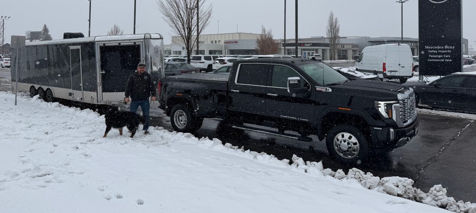 Adam and his dog beside the black GMC Denali and the enclosed triple-axle trailer in a snowy dealership lot