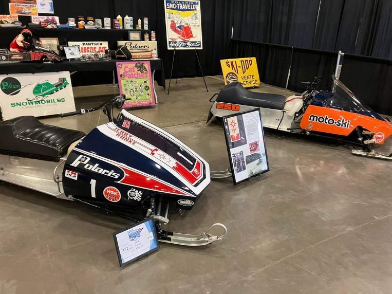 Vintage Polaris race sled with Jerry Bunke graphics and an orange Moto-Ski on display at a snowmobile show
